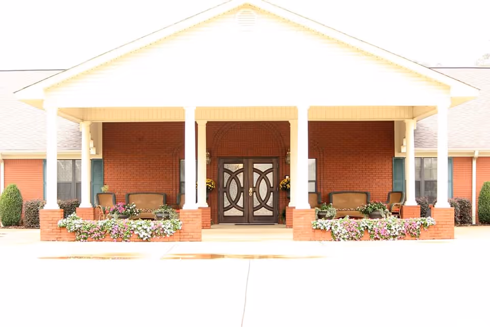 Front exterior view of Southern Magnolia Golden facility showing a covered entrance with white columns, brick walls, decorative double doors, seating areas with chairs and small tables, and flower beds with colorful flowers.