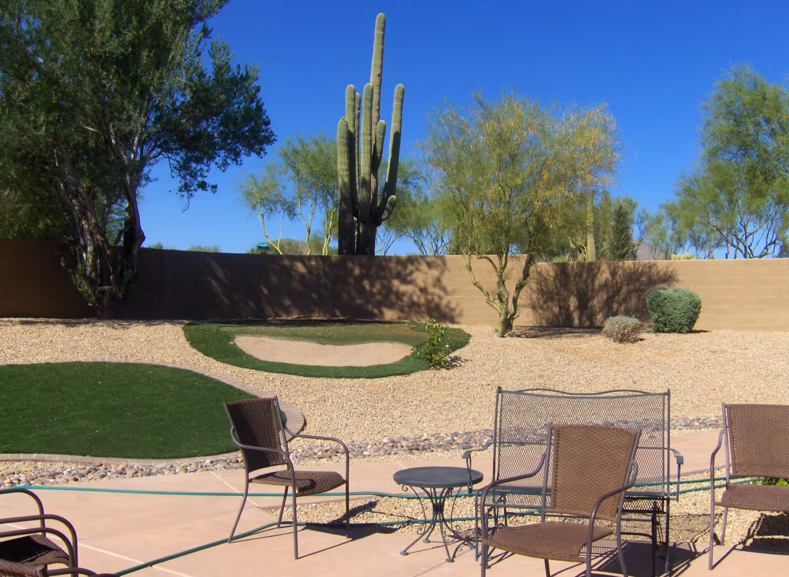 Outdoor patio area with metal chairs and a small round table on a concrete surface. In the background, there is a landscaped yard with gravel, green artificial turf patches, desert plants, trees, and a tall cactus against a clear blue sky.