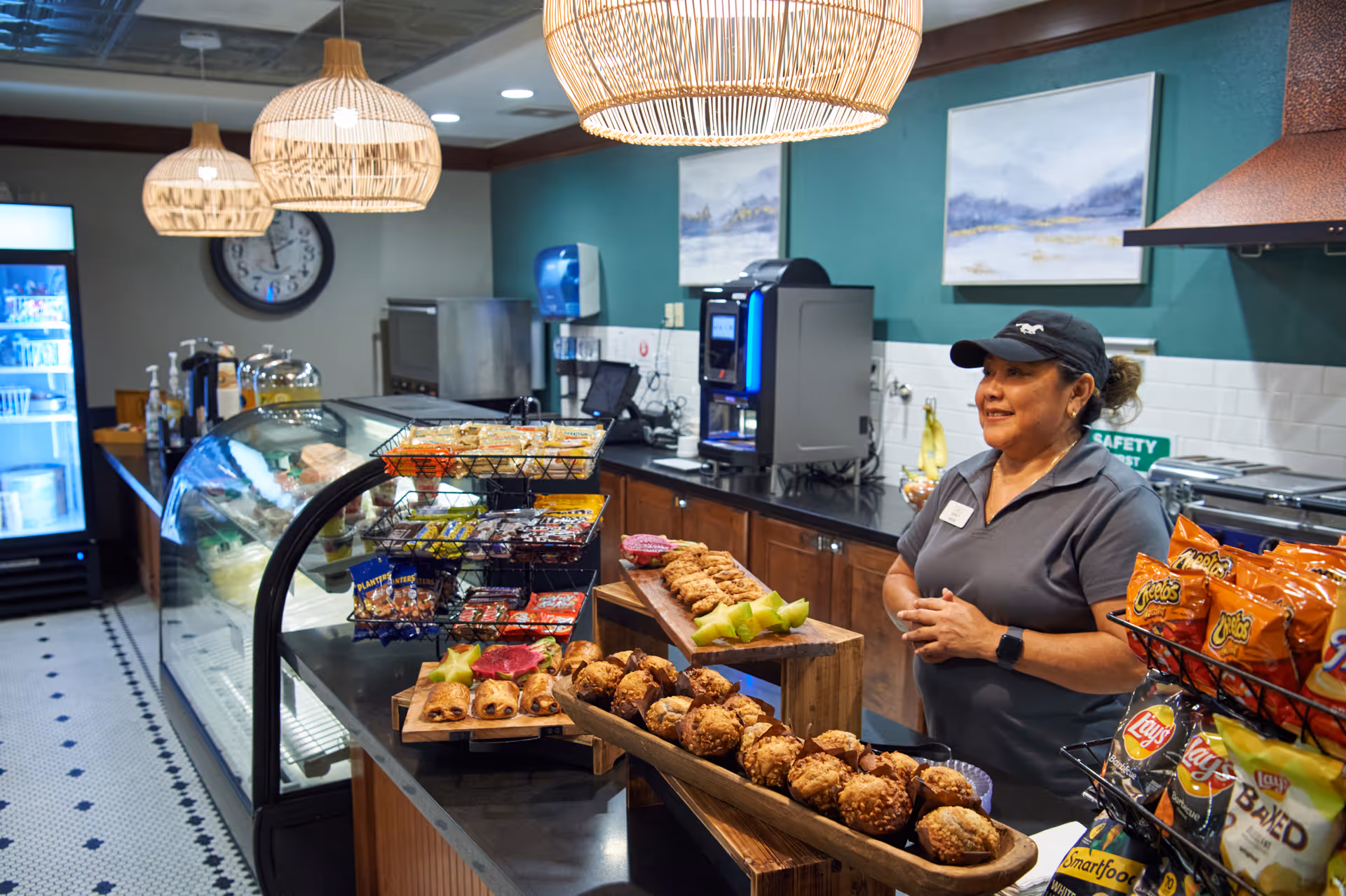 A staff member stands behind a café counter filled with pastries, fruit, snacks and a refrigerated display in an indoor dining area.
