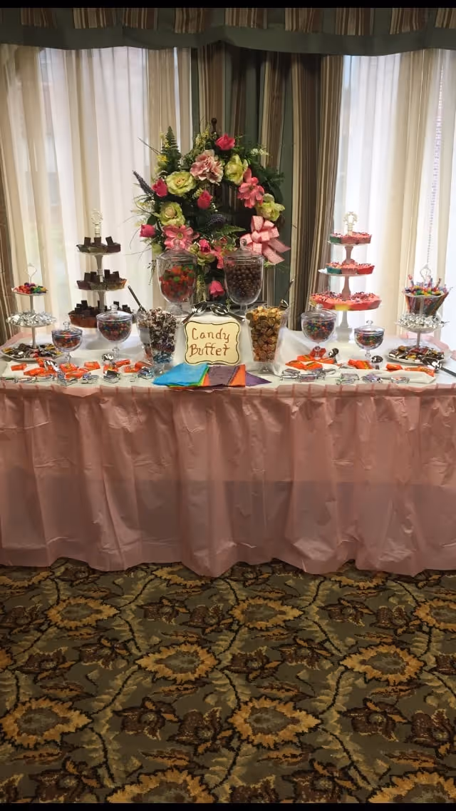 A candy buffet table decorated with a pink tablecloth and a floral arrangement in the center. The table holds various jars and trays filled with different types of candies, including chocolates, gummies, and wrapped sweets. Behind the table are large windows with sheer curtains and striped drapes.