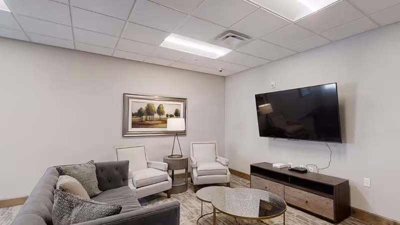 A small lounge with a sofa, two armchairs, a round coffee table, wall-mounted TV, and framed artwork.
