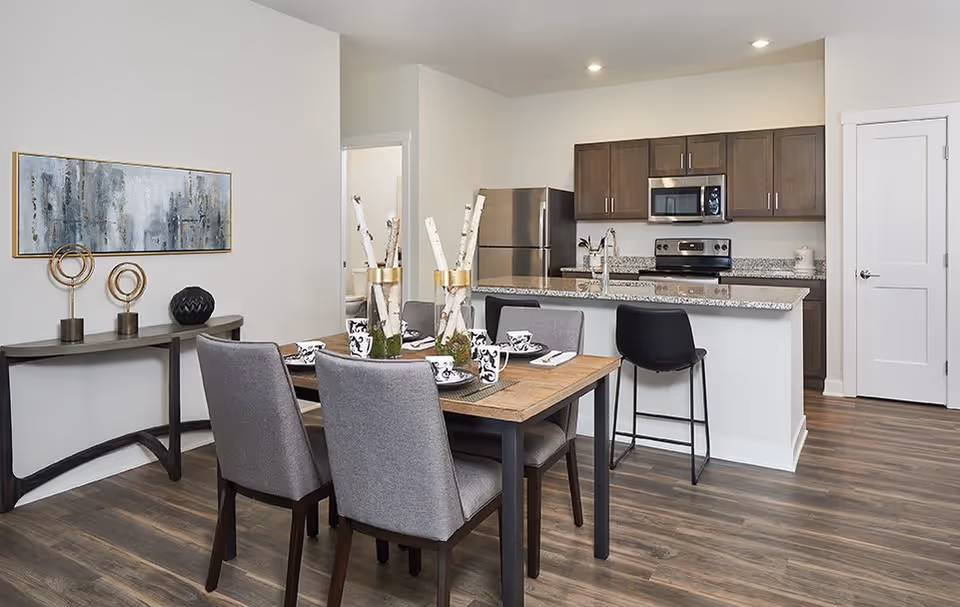 A modern dining area and kitchen in a senior living facility. The dining table is set with four gray upholstered chairs and decorative tableware. The kitchen features granite countertops, stainless steel appliances including a refrigerator, stove, and microwave, and dark wood cabinets. The floor is wood laminate, and there is a decorative console table with art and sculptures against the wall.