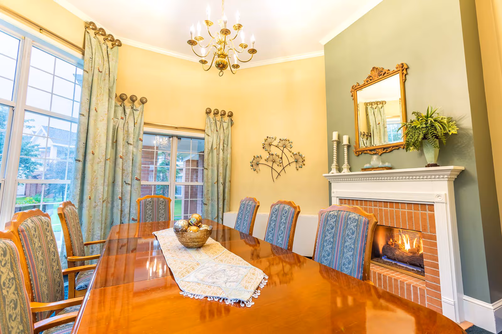 A formal dining room with a polished wooden table surrounded by upholstered chairs. The room features large windows with patterned curtains, a lit brick fireplace with a white mantel, decorative candles, a plant, and a gold-framed mirror above the fireplace. A chandelier hangs from the ceiling, and a decorative wall art piece is mounted on the wall.