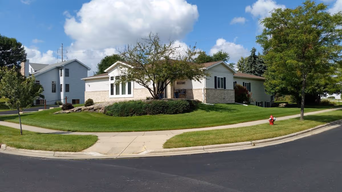 Exterior view of a single-story building with white siding and stone accents, surrounded by green grass, trees, and a sidewalk. A red fire hydrant is visible near the curb, and the sky is partly cloudy.
