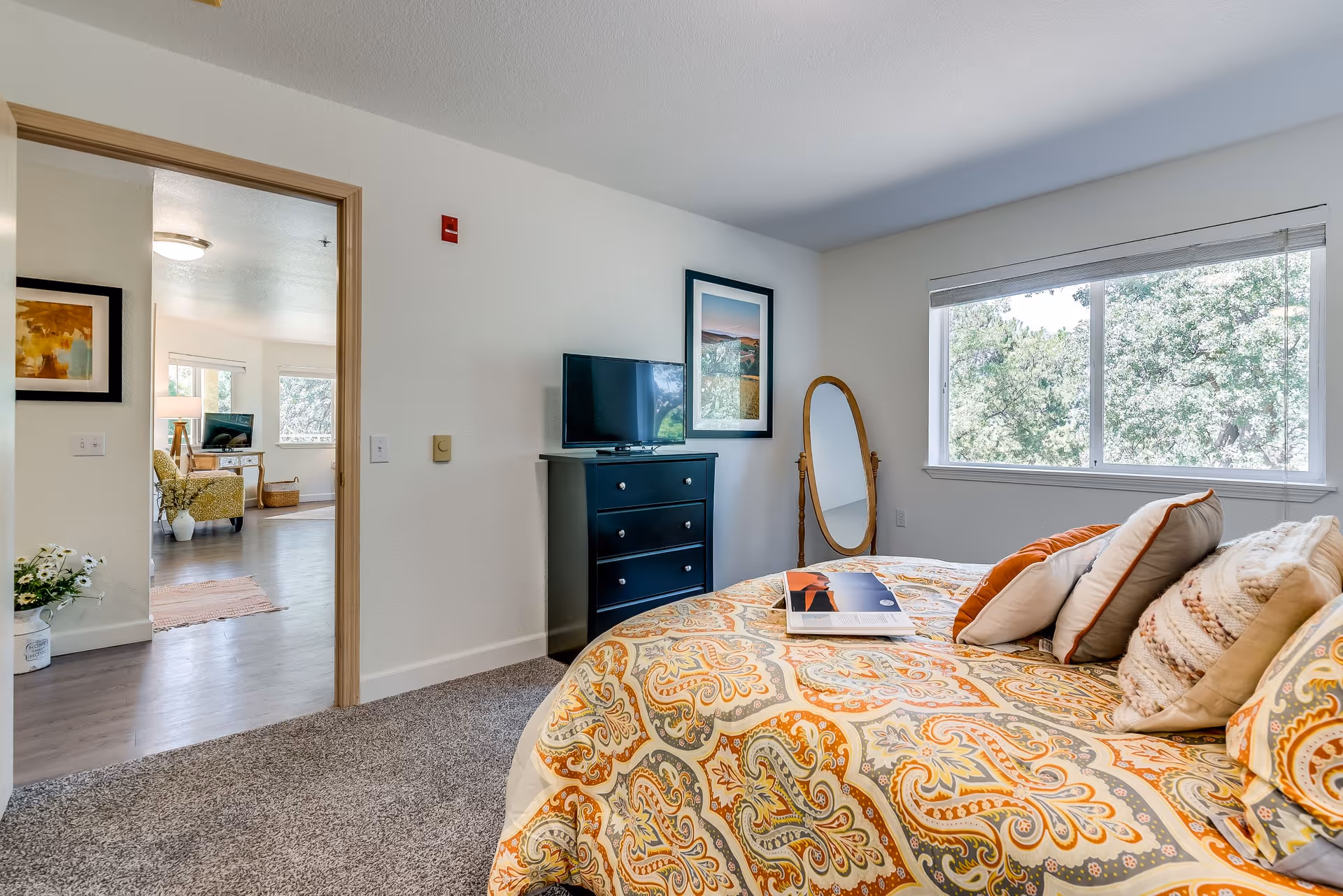A bright bedroom in a senior living facility with a patterned bedspread and multiple pillows on the bed. A black dresser with a TV on top and a framed picture above it is against the wall. A full-length wooden framed mirror stands near a large window showing green trees outside. The bedroom opens into a living area with chairs, a TV, and large windows.