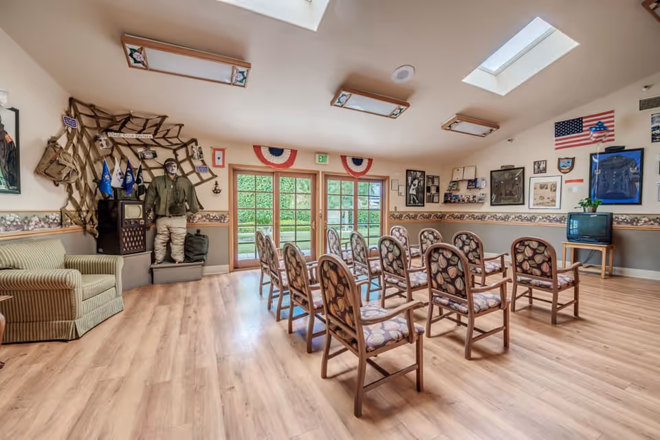 A memory care facility room with rows of wooden chairs featuring patterned cushions arranged facing a small TV on a stand. The room has light wood flooring, beige walls with a decorative border, and patriotic decorations including American flags and red, white, and blue bunting above glass double doors leading outside. There is a mannequin dressed in military attire standing next to an old-fashioned television and military memorabilia displayed on the walls.