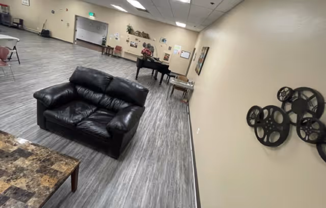 A spacious room with gray wood flooring and beige walls, featuring a black leather loveseat, a marble-patterned coffee table, a grand piano with a floral arrangement on top, and wall decorations including film reel art. There are also chairs and tables along the walls, and a doorway leading to another area.