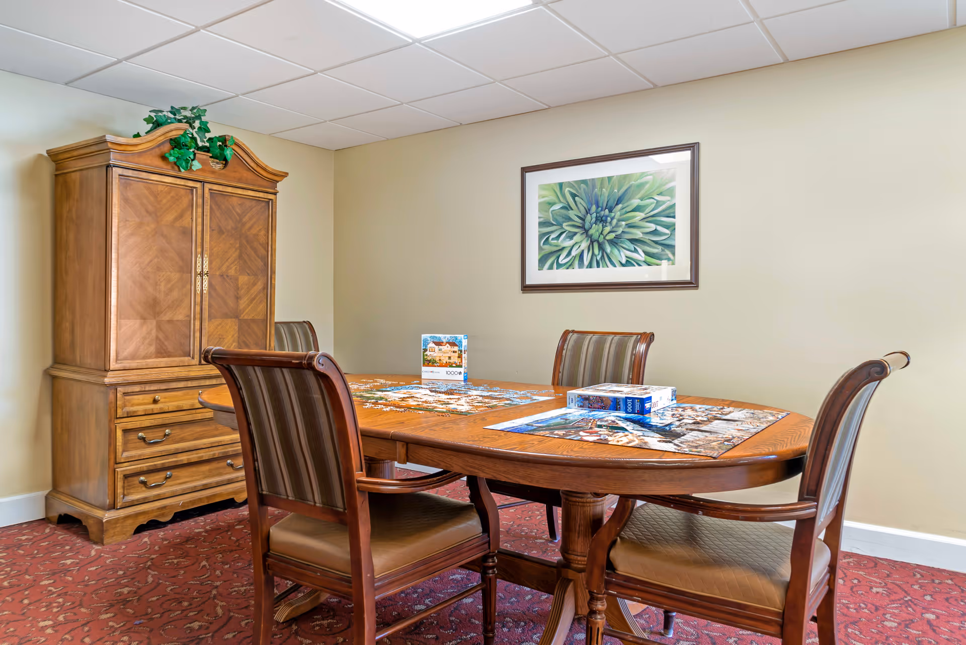 A room with a wooden oval table and four chairs with striped upholstery. On the table, there are two partially completed jigsaw puzzles and puzzle boxes. Behind the table is a wooden cabinet with drawers and a decorative plant on top. A framed picture of a green flower hangs on the beige wall. The floor is covered with a red patterned carpet, and the ceiling has white tiles.