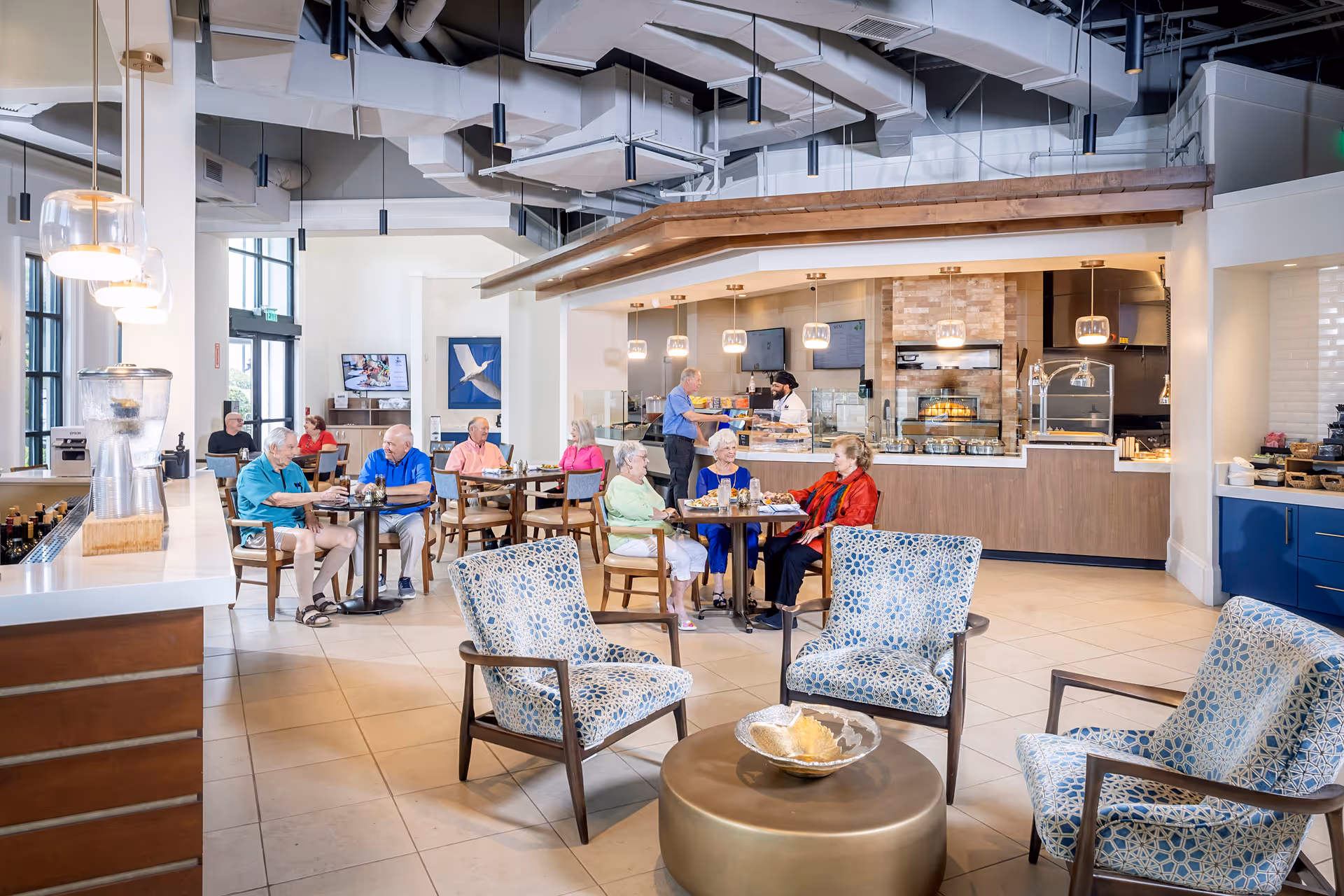 Bright communal dining/café area with several older adults seated at tables and staff serving behind a counter.