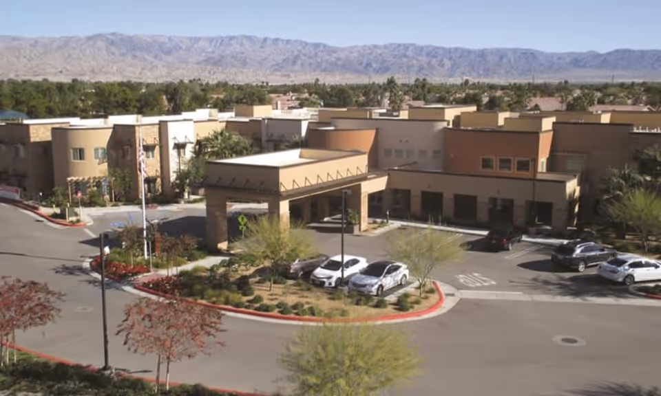 Exterior view of The Palms At La Quinta Senior Living facility showing a large building with a covered entrance, several parked cars, landscaped areas with trees and shrubs, and mountains in the background under a clear sky.