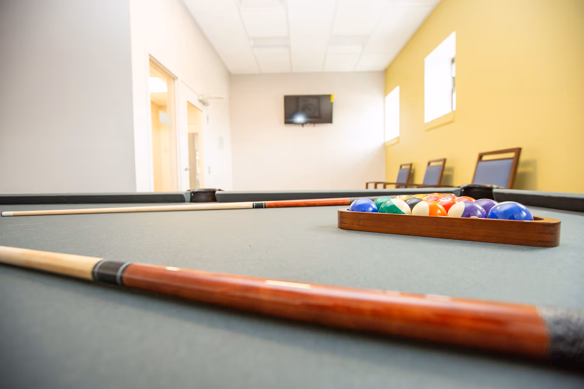 Close-up view of a pool table with a rack of billiard balls and two pool cues resting on the table in a well-lit room with yellow and white walls, chairs along the wall, and a wall-mounted TV in the background.