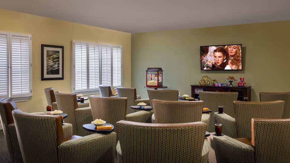 A cozy common room with multiple patterned armchairs arranged in rows facing a wall-mounted television showing a movie scene. Each chair has a small side table with snacks and drinks. There is a popcorn machine on a stand near the TV, and the room has light green walls and windows with white shutters.