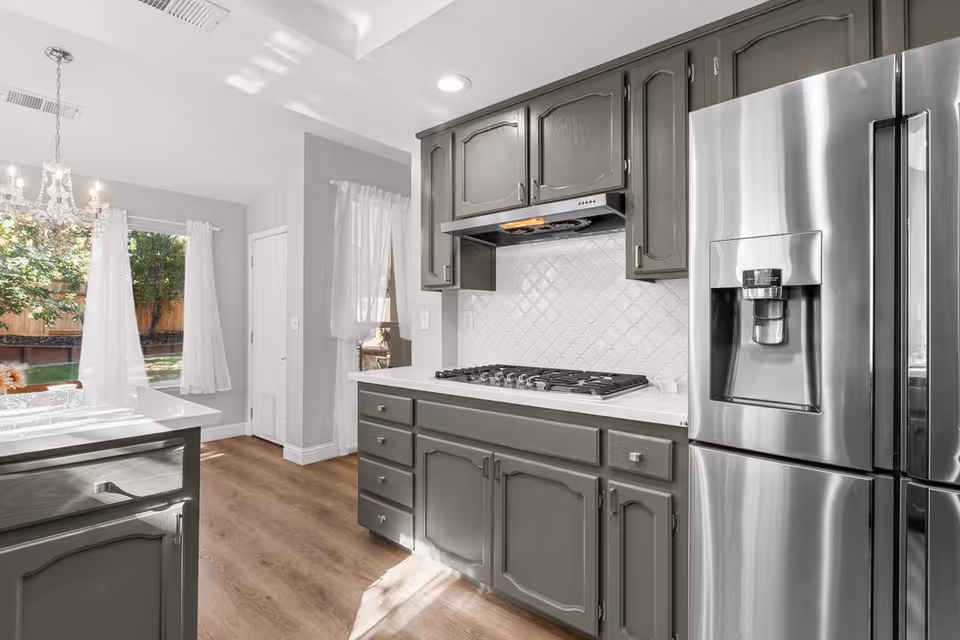Modern kitchen with gray cabinets, a stainless steel refrigerator with water and ice dispenser, a gas stove with a range hood, white tiled backsplash, and a wooden floor. There is a dining area visible in the background with a chandelier and large windows with white curtains letting in natural light.