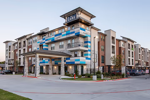 Exterior view of The Luxe at Cedar Hill, a modern multi-story senior living facility with a covered entrance, stone and brick facade, and blue and white decorative panels. The building is surrounded by a driveway and parking area with some landscaping.