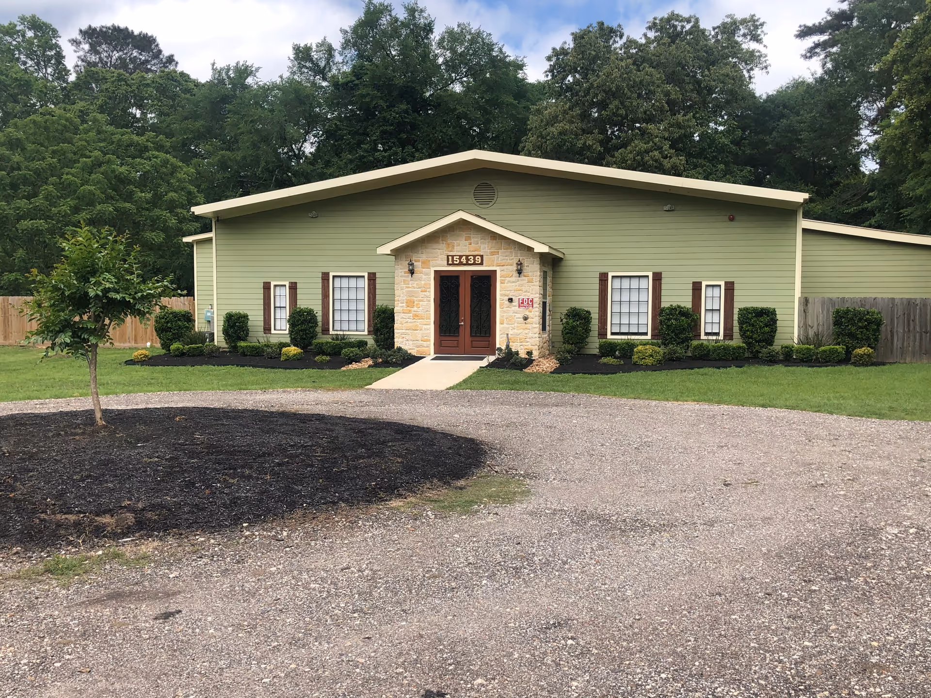 Single-story green building with a central stone entrance, double doors, and landscaped front lawn and driveway.