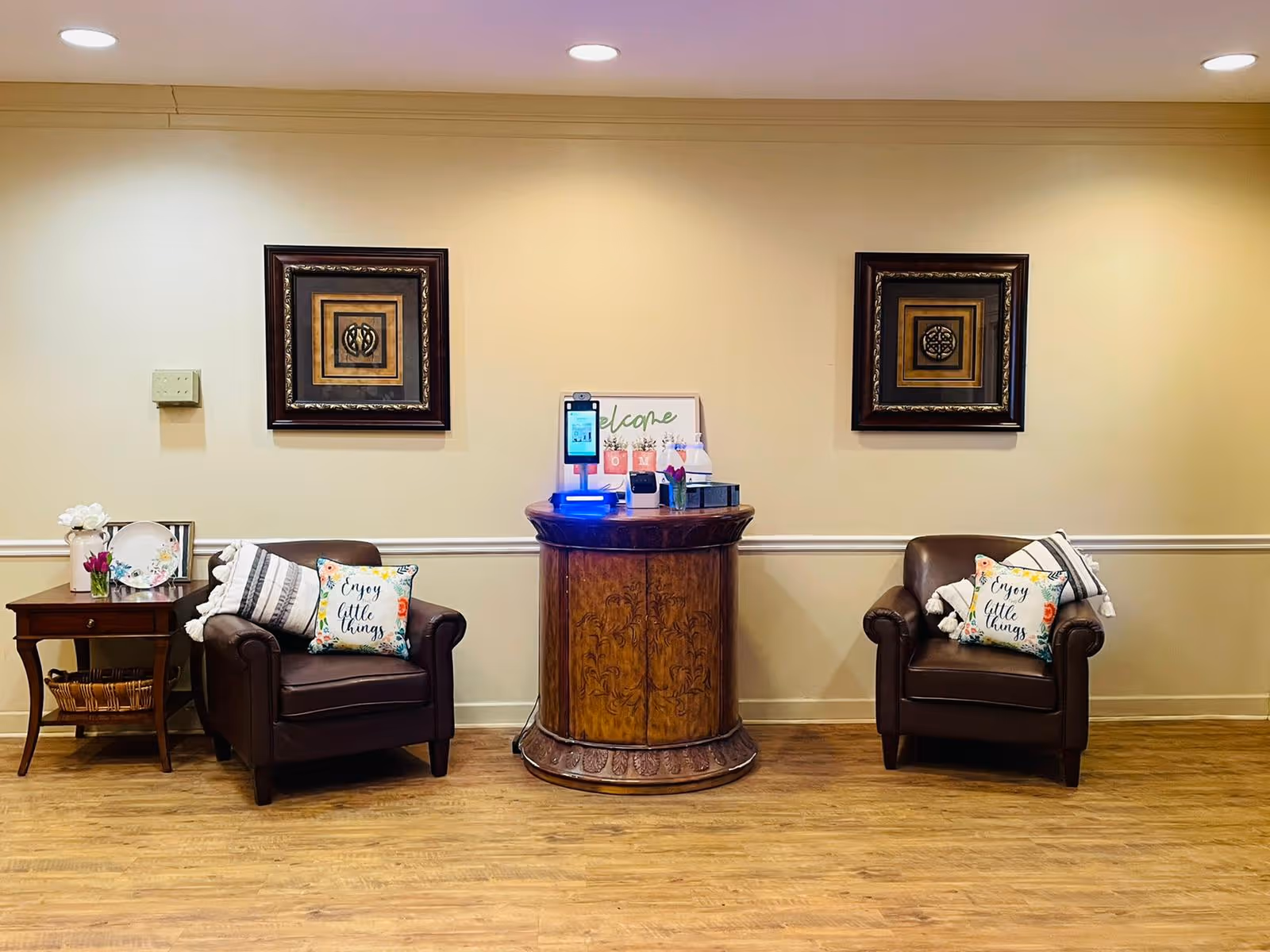 A cozy seating area with two brown leather armchairs, each adorned with a decorative pillow that says 'Enjoy little things' and a folded blanket. Between the chairs is a round wooden table with a welcome sign, hand sanitizer, and other small items. To the left of the left chair is a small wooden side table with a flower vase and decorative plates. The wall behind has two framed square art pieces and is painted a light beige color. The floor is wood.