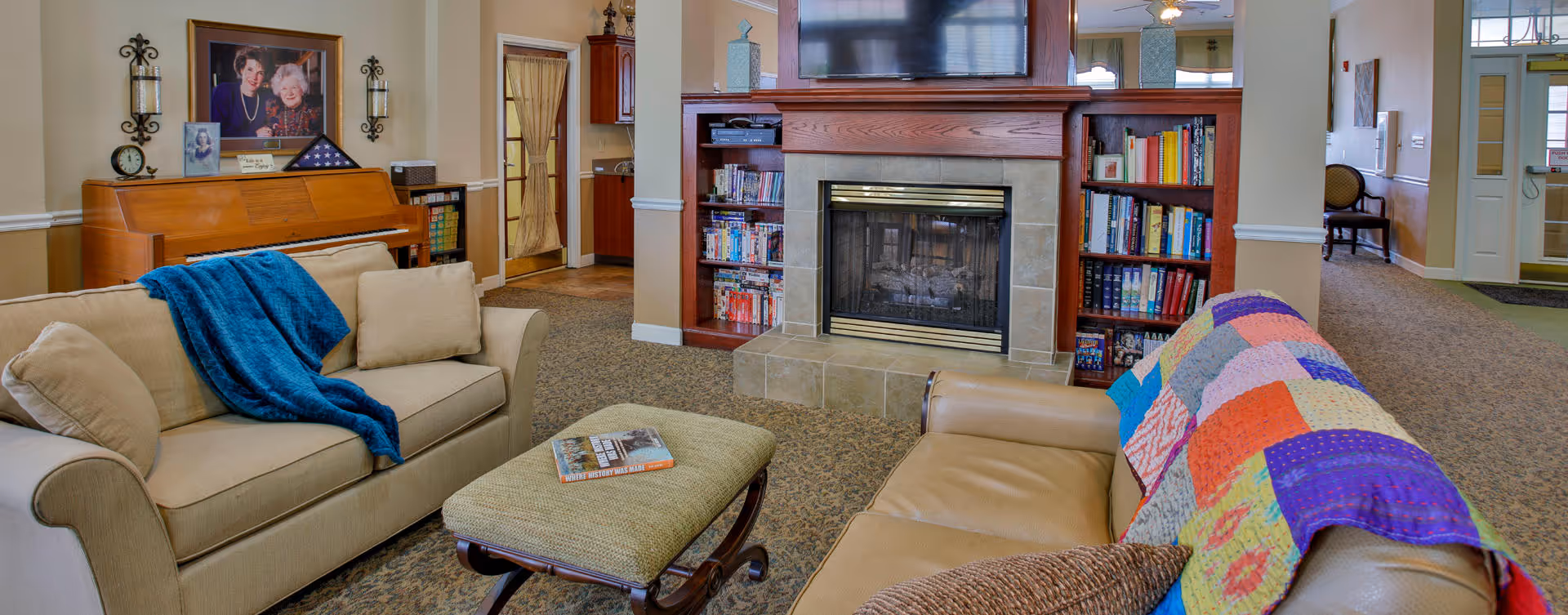 Cozy common living room with sofas, an ottoman, a quilt, a fireplace flanked by bookshelves, a TV, and a piano in the background.
