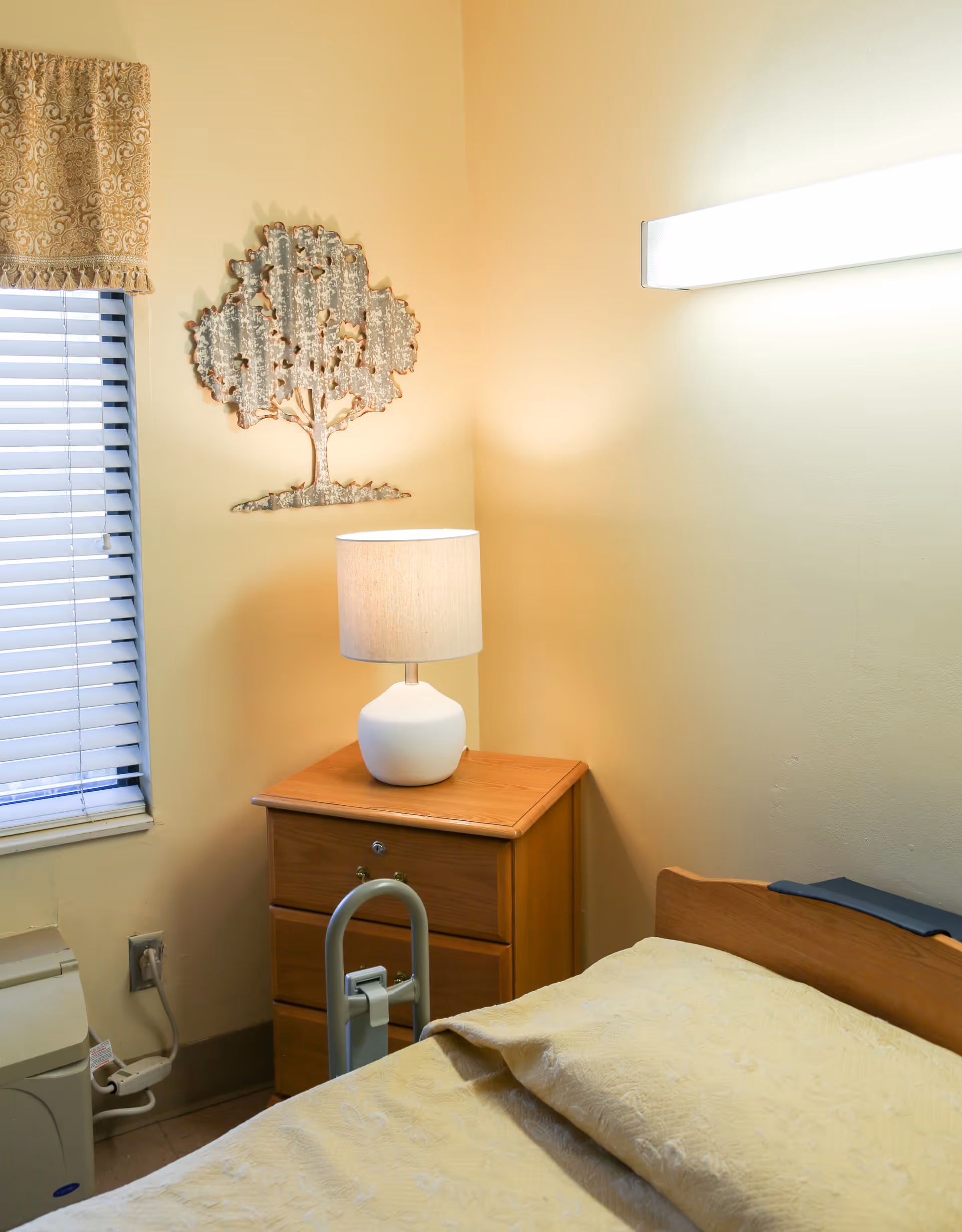 Simple bedroom corner with a hospital-style bed, wooden nightstand topped by a lamp, wall art on the wall, and a window with blinds.