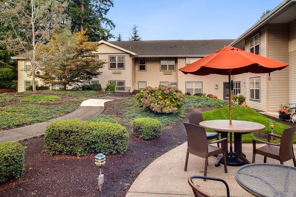 Outdoor patio area at Canfield Place featuring a round table with a large red umbrella and several chairs. The patio is surrounded by landscaped bushes, a curved concrete walkway, and a two-story beige building with multiple windows in the background. Trees and greenery are visible under a clear blue sky.