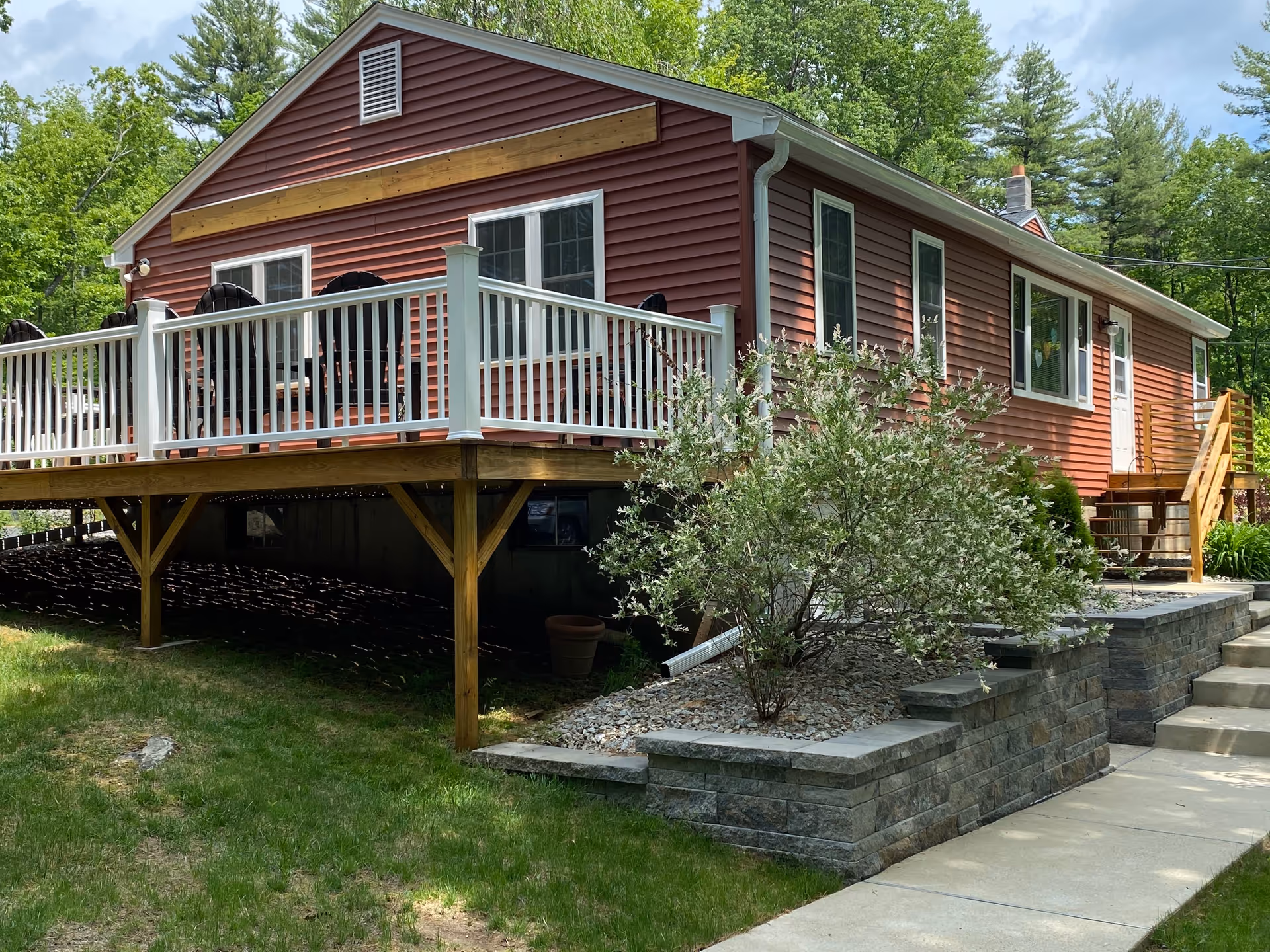 Exterior view of a single-story red house with white trim and a wooden deck with white railings. The house is surrounded by green trees and shrubs, with a stone retaining wall and concrete walkway leading to the entrance.