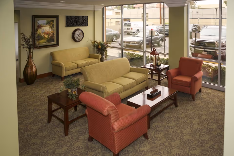 A cozy living room area with two beige sofas, two red armchairs, a wooden coffee table, and side tables. The room has large windows showing parked cars outside, a wall clock, a framed painting, and decorative plants.