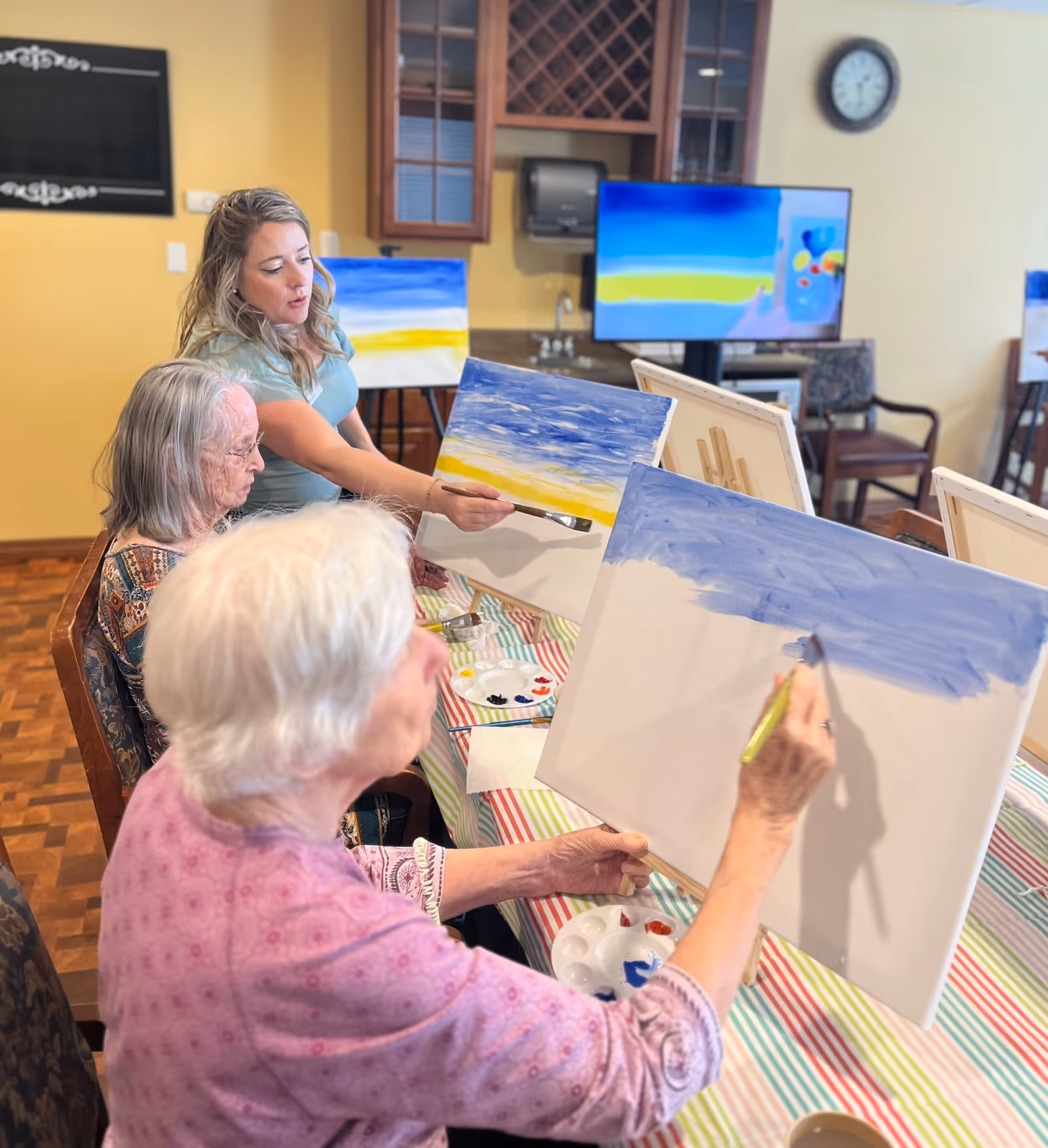 Two elderly women and a younger woman seated at a table engaged in a painting activity. The elderly women are painting on canvases with blue and yellow colors, while the younger woman appears to be instructing or assisting them. The room has a warm yellow wall, a clock, a TV screen displaying a similar painting, and wooden cabinets in the background.