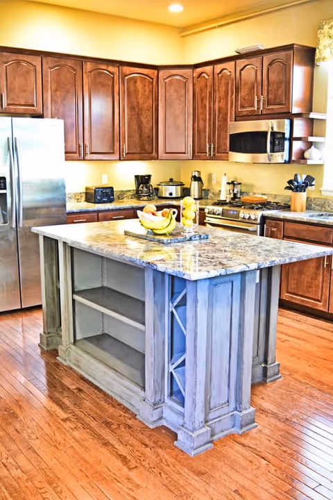 A bright kitchen with wooden cabinets, stainless steel refrigerator, microwave, and stove. There is a kitchen island with a marble countertop in the center, featuring open shelves and a decorative wine rack. Various kitchen appliances and utensils are placed on the countertops, and a bowl of fruit sits on the island.