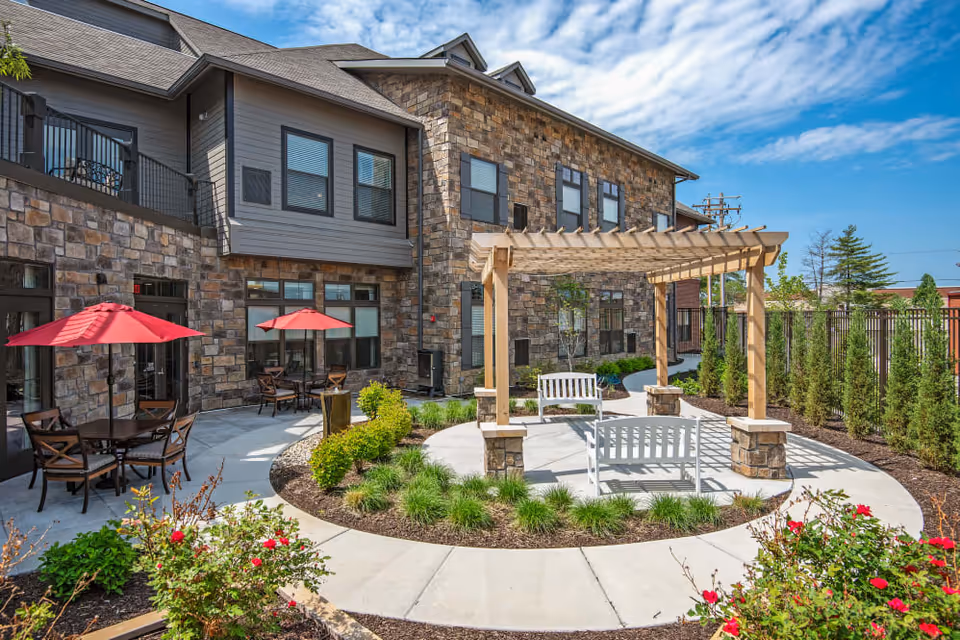 Outdoor courtyard with a wooden pergola, white benches, patio tables with red umbrellas, landscaping, and a stone-clad building in the background.
