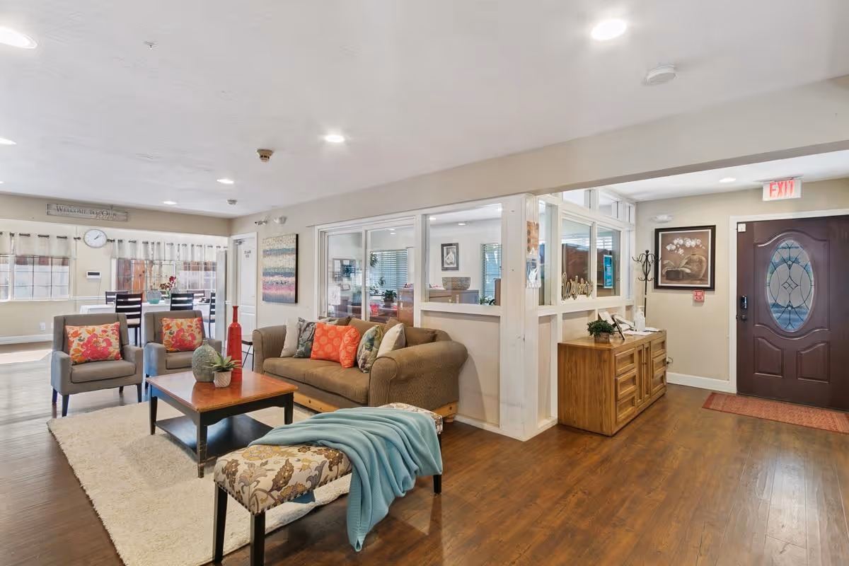 A bright and welcoming senior center common area with a brown sofa adorned with colorful pillows, two gray armchairs with floral cushions, a wooden coffee table with a plant, and a patterned bench with a blue throw. The room has wooden flooring, white walls, and large windows allowing natural light. In the background, there is a dining area with a table and chairs, a wooden sideboard with decor, and a dark wooden door with a decorative glass panel.