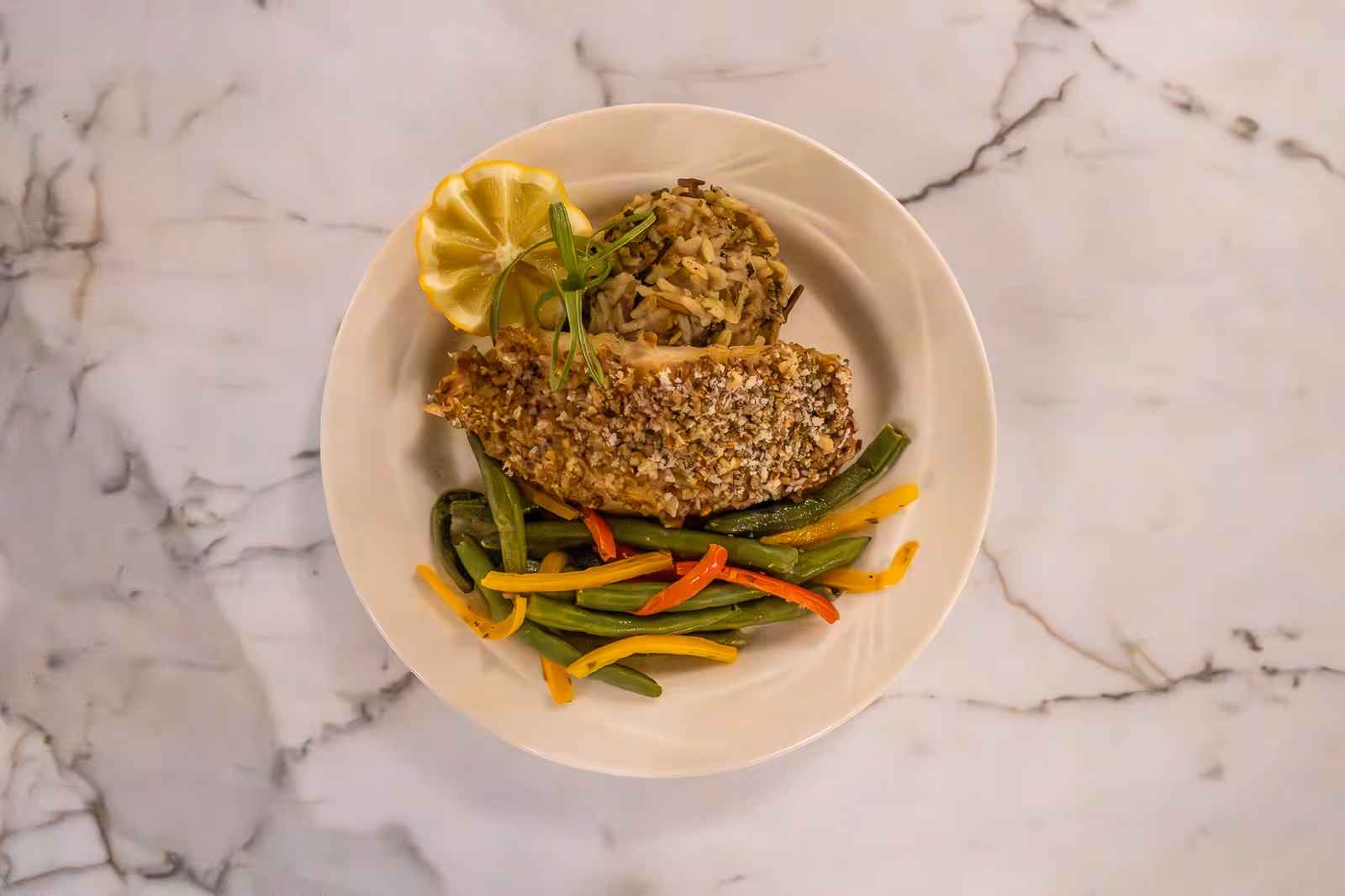 A white plate with a serving of breaded fish, a portion of mixed wild rice, green beans, and sliced yellow and red bell peppers, garnished with a lemon wedge and a sprig of green herb, placed on a marble surface.