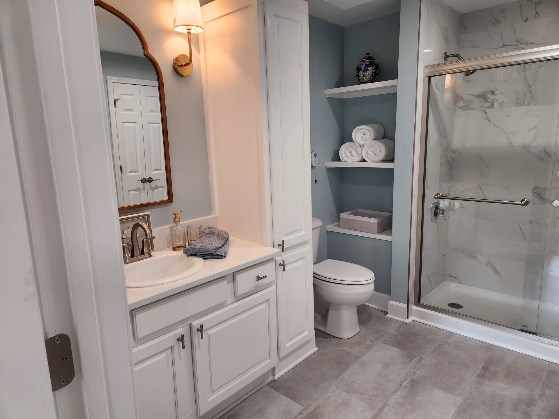 Clean bathroom with a white vanity and sink, toilet with open shelving holding rolled towels, and a glass-enclosed marble shower.