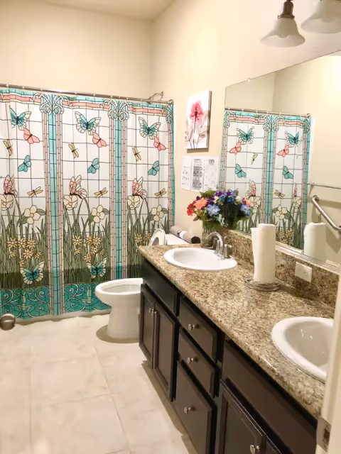 Bathroom with double sinks, granite counter, butterfly-pattern shower curtain, and flowers.