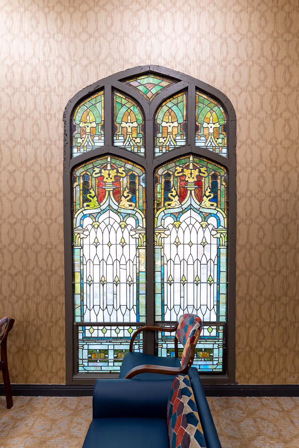 Tall arched stained-glass window set in a patterned wallpapered wall with upholstered chairs in front.