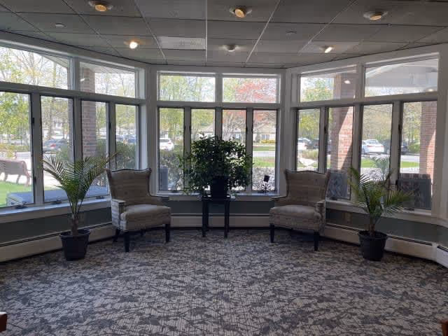 A bright seating area with large windows overlooking a parking lot and trees outside. The room features two upholstered armchairs separated by a small black table with a potted plant on it. There are two additional potted plants on either side near the windows. The floor is carpeted with a patterned design, and the ceiling has recessed lighting.