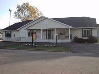 Single-story light-colored assisted living building with a covered front porch, columns, and a small lawn with a sign.