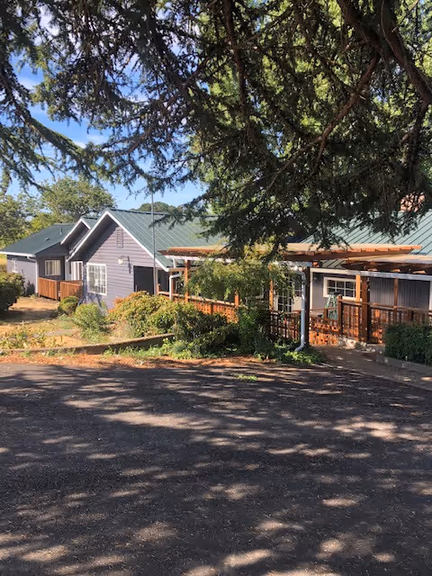 Single-story retirement village building with green metal roofs, a wooden porch and railings, and trees casting shadows across the paved foreground.