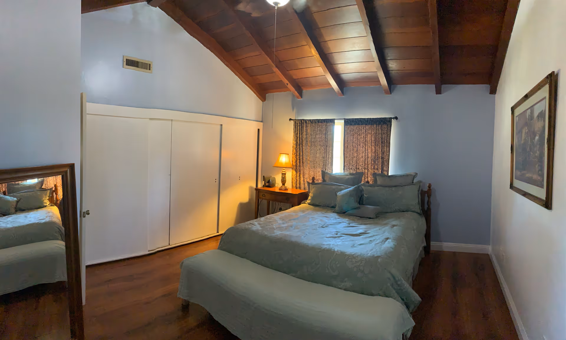 A cozy bedroom with a bed, nightstand, window, and exposed wooden ceiling beams.