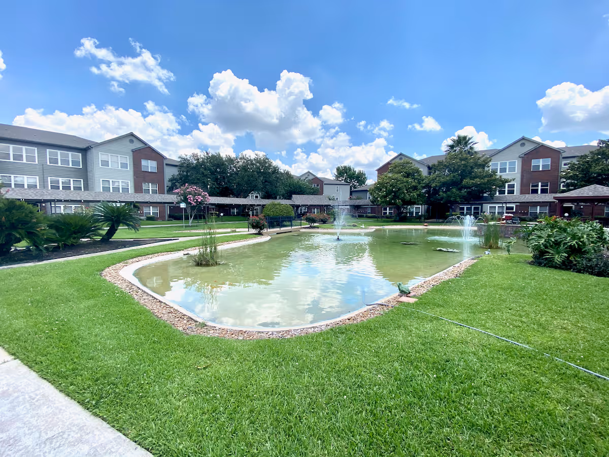 Outdoor view of a senior living facility with a large pond featuring multiple water fountains surrounded by well-maintained green grass and landscaped plants. The multi-story residential buildings are visible in the background under a partly cloudy blue sky.