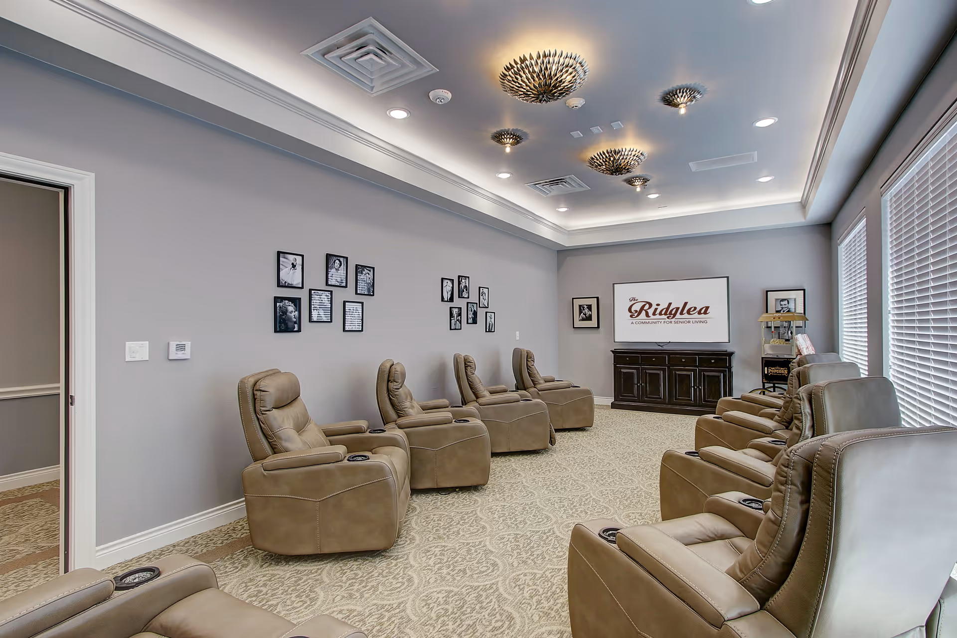 A senior living facility lounge with two rows of comfortable beige recliner chairs facing a large flat-screen TV mounted on a dark wooden cabinet. The walls are painted light gray and decorated with framed black and white photos. The ceiling features modern, decorative light fixtures and recessed lighting. Large windows with white blinds allow natural light into the room.