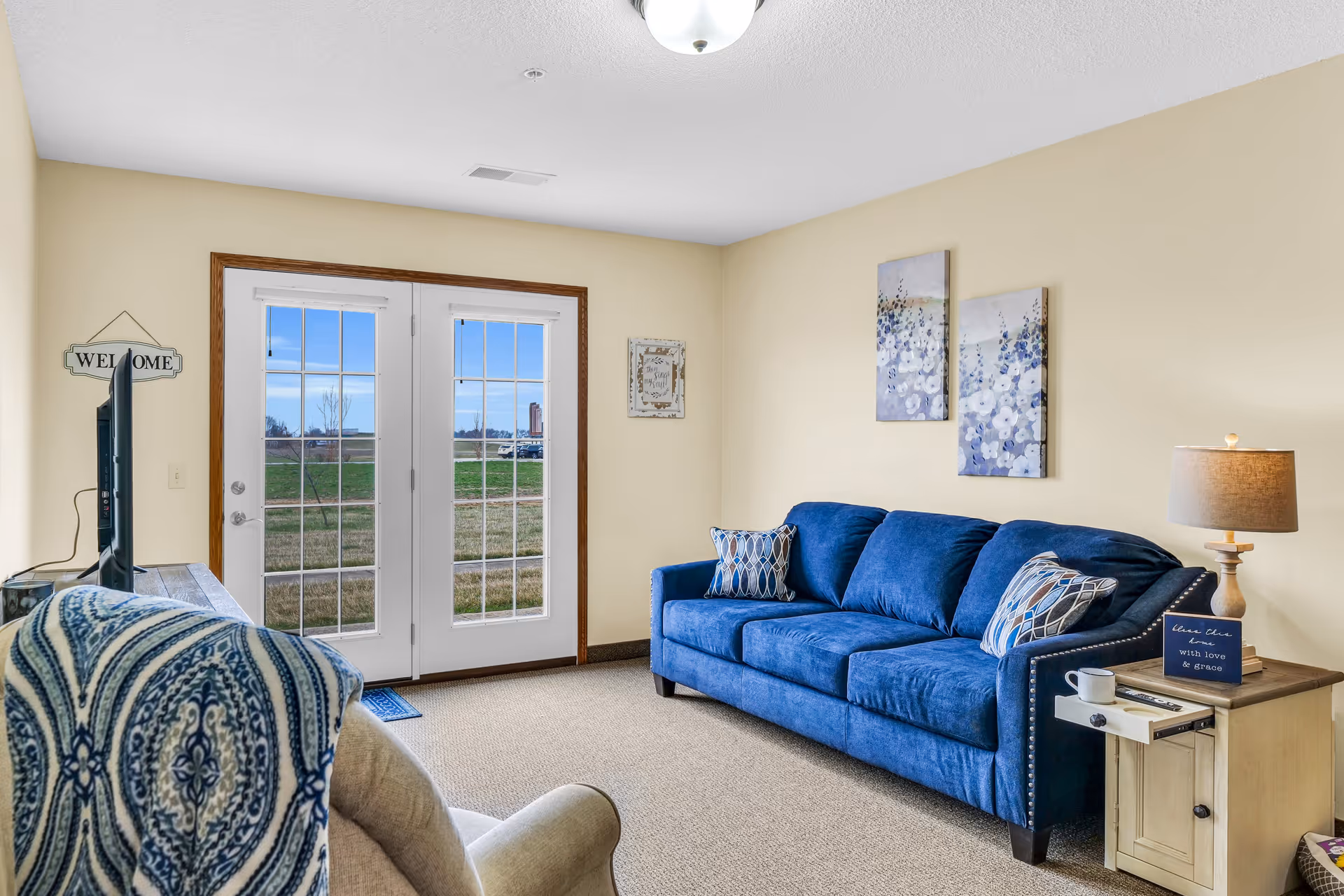 A cozy living room with a blue couch adorned with patterned pillows, a beige armchair with a blue and white patterned throw, a wooden side table with a lamp and a small sign, and double glass doors leading outside to a grassy area under a clear blue sky.