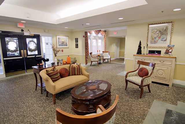 A cozy senior living facility common area with beige walls and patterned carpet. The room features a beige loveseat with striped and round pillows, a wooden coffee table with a glass top, and two wooden chairs with patterned cushions. There is a sideboard with a small Christmas tree and framed artwork above it. Double doors with wreaths are visible on the left, and a small seating area with two chairs and a table is in the background near windows with red and white curtains.