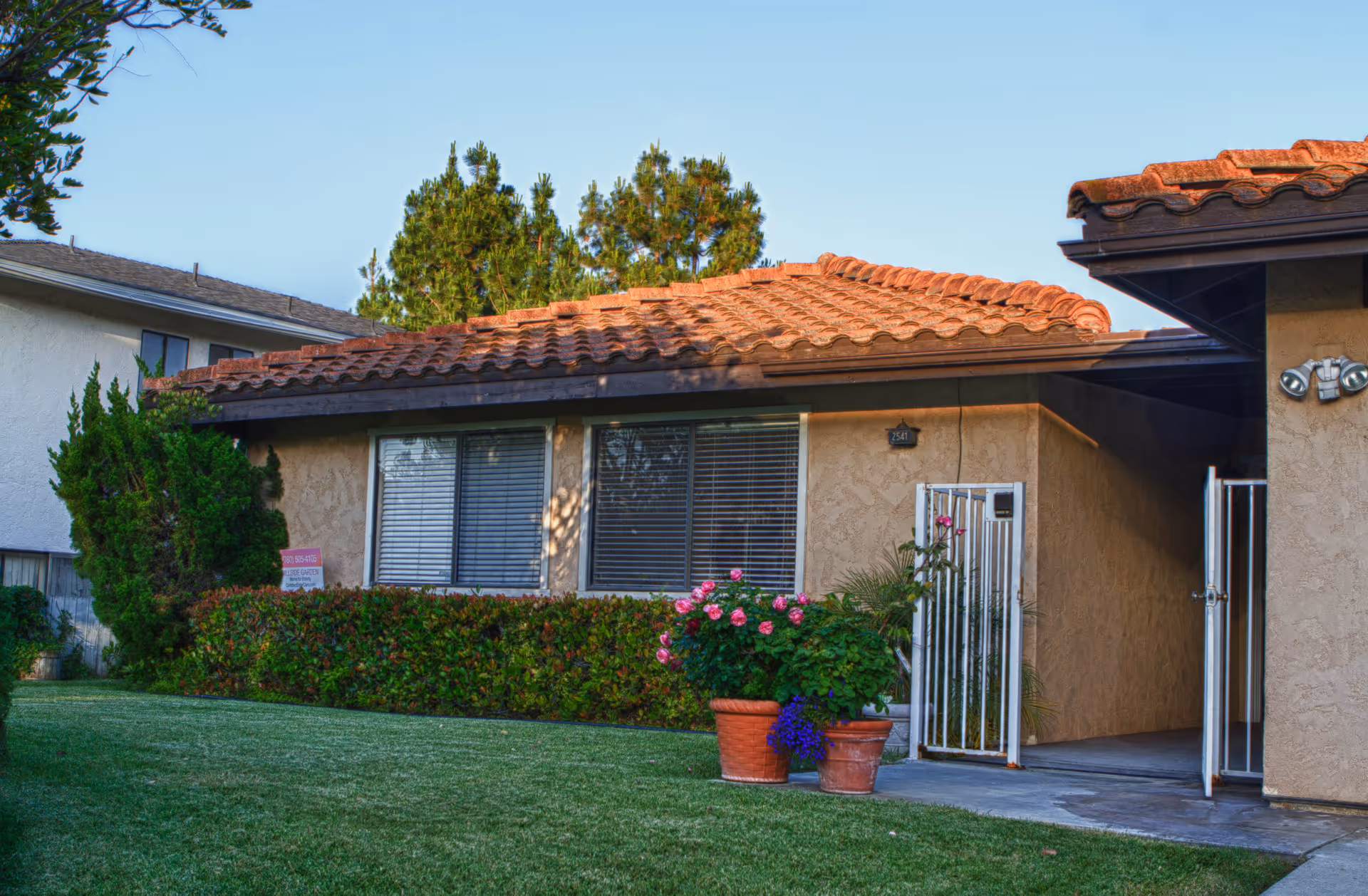 Exterior view of a single-story residential building with a tiled roof, beige stucco walls, and large windows with blinds. There is a small gated entrance with potted plants featuring pink and purple flowers in front. The lawn is well-maintained with green grass and trimmed bushes, and there are trees in the background under a clear sky.