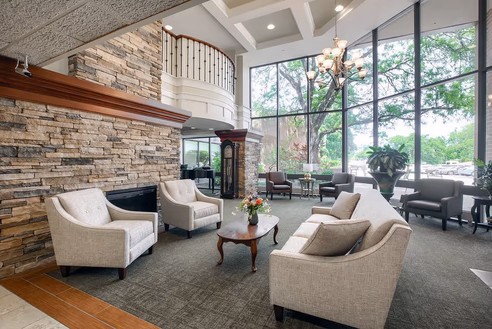 A spacious living room area with large floor-to-ceiling windows showing trees and a parking lot outside. The room features a stone fireplace, beige upholstered sofa and armchairs, a wooden coffee table with a flower arrangement, a grandfather clock, and a chandelier hanging from a coffered ceiling.