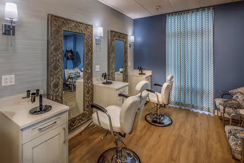 Interior of a salon room with two white salon chairs in front of two large ornate mirrors mounted on a light gray wall. Each station has a small white cabinet with a black faucet and sink. The floor is wood, and there is a blue wall with a patterned curtain covering a window. Two floral upholstered chairs are visible on the right side.