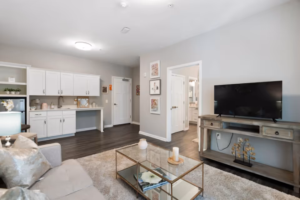 A modern living room area with a light gray sofa adorned with metallic pillows, a glass coffee table with a candle and magazines, and a wooden TV stand holding a flat-screen television. The room features white cabinetry with a small sink and countertop, dark wood flooring, and light gray walls. There is a doorway leading to another room and framed artwork on the wall.