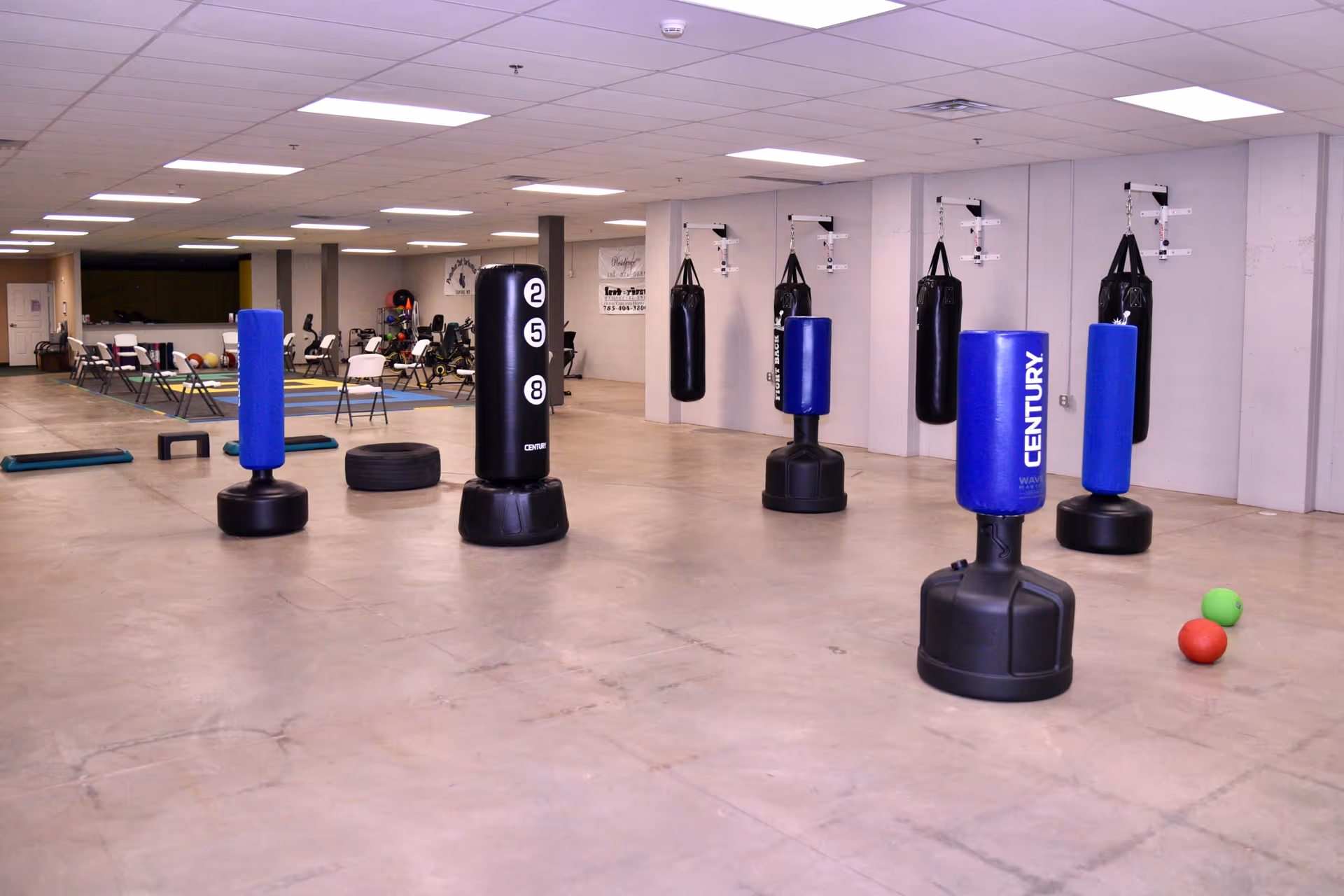 A spacious indoor fitness area with several punching bags, including freestanding blue and black bags and hanging black bags mounted on the wall. In the background, there are chairs arranged around exercise mats and various fitness equipment.