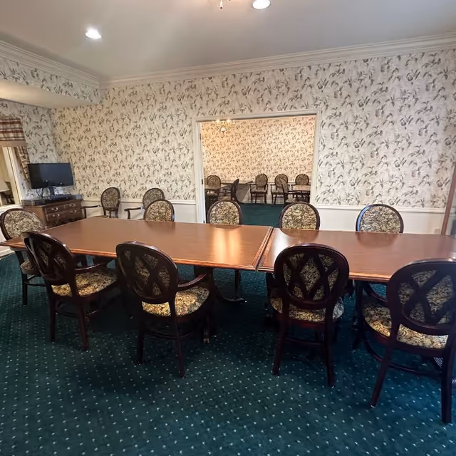 A dining room with two long wooden tables pushed together, surrounded by upholstered chairs with patterned fabric. The room has floral wallpaper, green carpet, and a small TV on a wooden dresser in the corner. Through an open doorway, another similar dining area with more chairs and tables is visible.