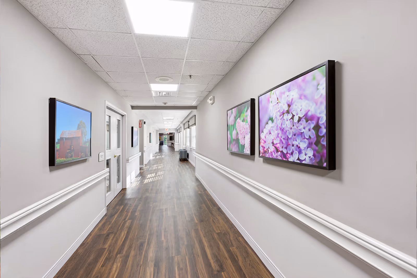 A long, well-lit hallway in a senior living facility with wood-style flooring and light gray walls. The hallway features framed floral and landscape artwork on the walls, white trim and handrails, and a ceiling with recessed lighting panels. Doors and windows line the hallway, and there is a small seating area visible in the distance.