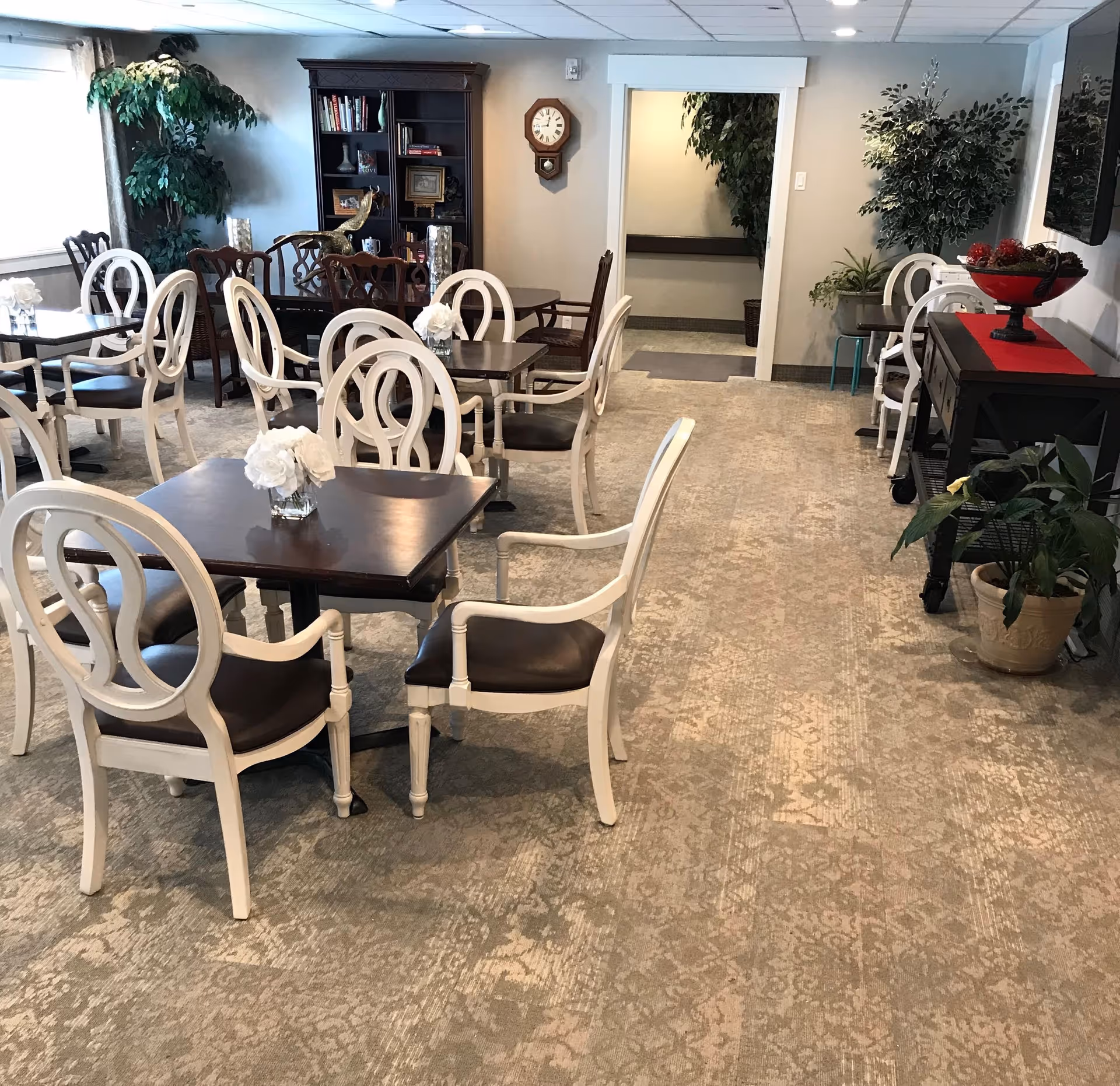 A dining room with several dark wood tables and white chairs with dark cushions. Each table has a small vase with white flowers. The room has a patterned carpet, potted plants, a wooden bookshelf with books and decorative items, a wall clock, and a sideboard with a red runner and a decorative bowl. There is a doorway leading to another room.