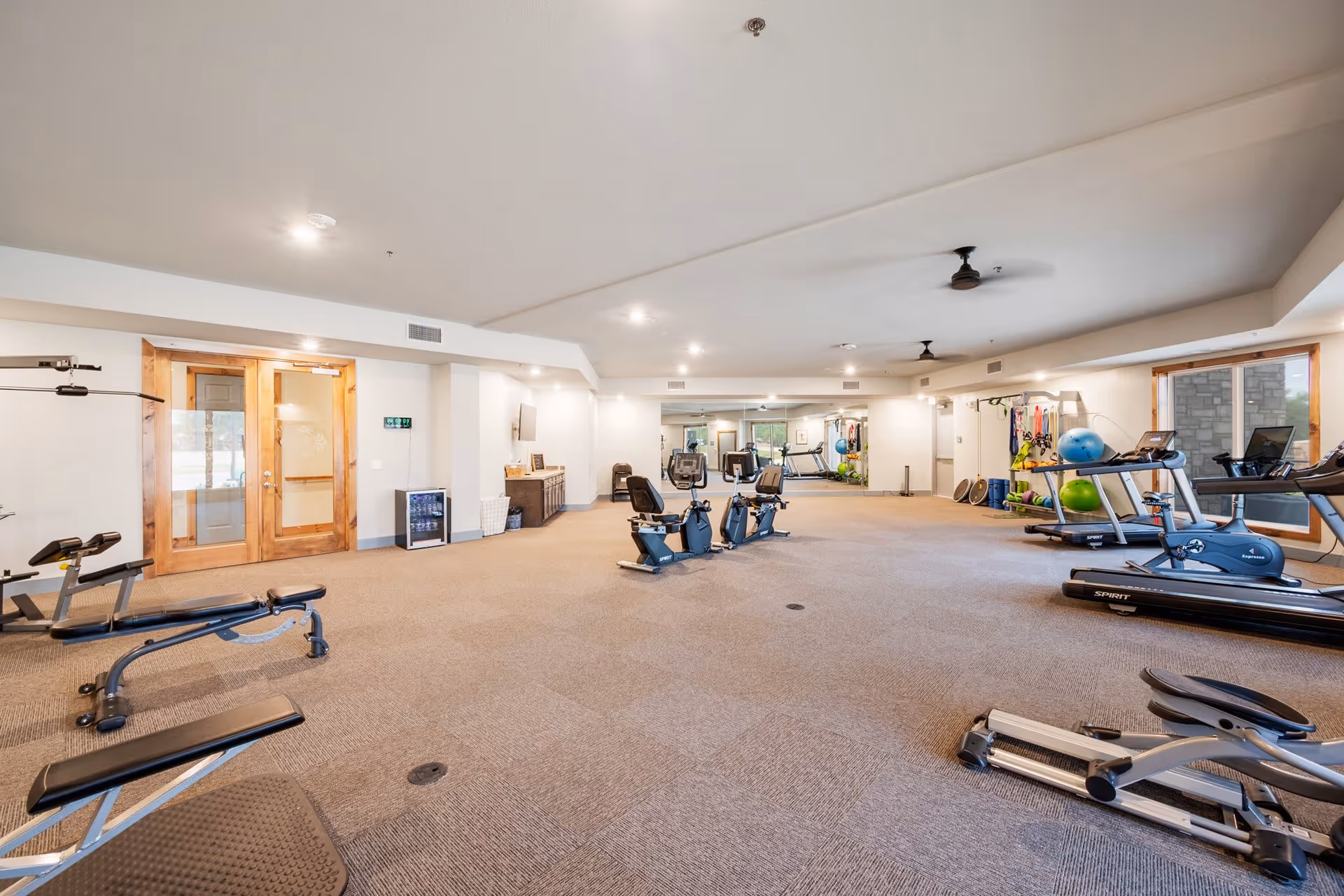 A spacious senior living facility fitness room with various exercise equipment including treadmills, stationary bikes, weight benches, and resistance bands. The room has beige carpeted floors, white walls, ceiling fans, and large windows letting in natural light. There is a wooden double door entrance and a wall-mounted TV with a small refrigerator below it.